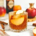 Glass of apple cinnamon whiskey cocktail garnished with apple slices and surrounded by fresh apples, cinnamon sticks, and a liquor bottle
