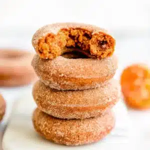 Stack of three cinnamon sugar pumpkin donuts with a bite taken out of the top one, set on a white surface