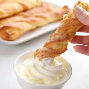 Hand holding a cinnamon sugar twist breadstick being dipped into a swirl of vanilla cream, with more breadsticks on a plate in the background