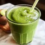 A thick, creamy green avocado smoothie in a glass with a polka-dot straw, placed on a marble surface with sliced avocado in the background.Green smoothie for weight loss