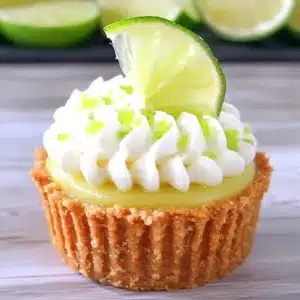 Mini key lime pie cupcake topped with piped whipped cream, lime zest, and a fresh lime wedge, in a graham cracker crust on a light wood surface.
