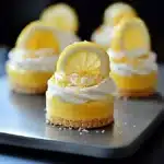 Small lemon cheesecake bites topped with whipped cream, golden sugar sprinkles, and fresh lemon slices on a dark tray.