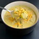 Close-up of creamy potato soup in a black bowl, topped with shredded cheddar cheese and chopped herbs