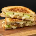 Golden grilled cheese sandwich with melted cheese, green apple slices, and caramelized onions on a wooden board.