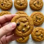 Hand holding a soft pumpkin chocolate chip cookie with more cookies arranged on a light surface in the background.